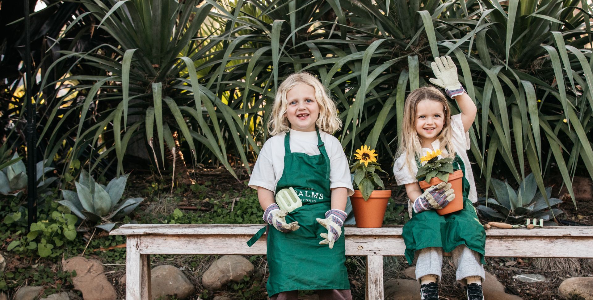 Two children in The Palms aprons sitting on a bench with sunflowers in a garden